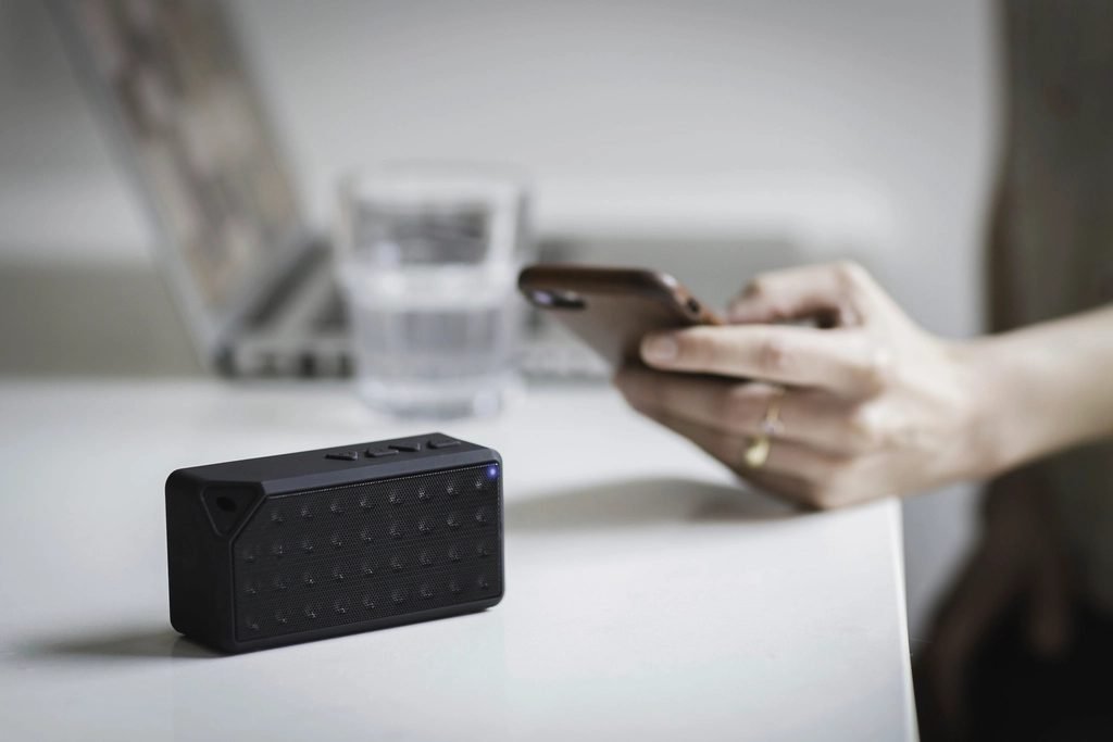 Hand holding a phone beside a small Bluetooth speaker on a white table