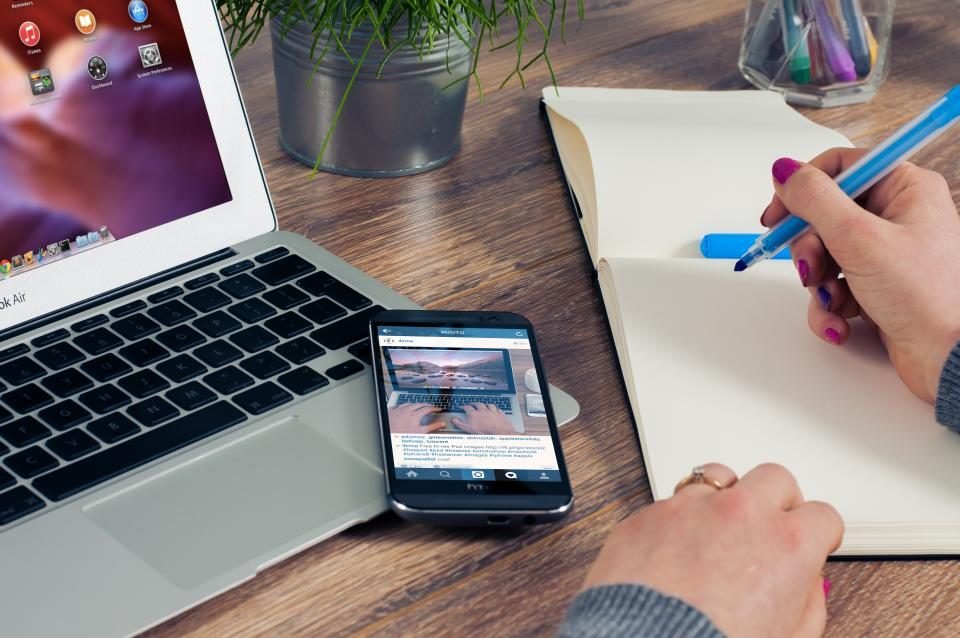Hands writing in a notebook next to a laptop and a phone