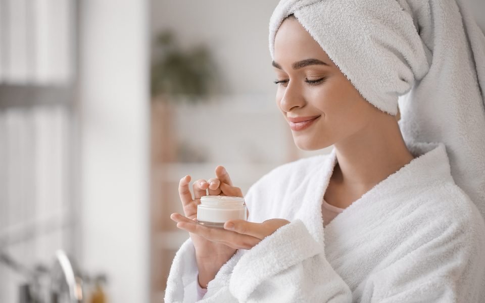 Woman in a white bathrobe and towel holding a jar of face cream